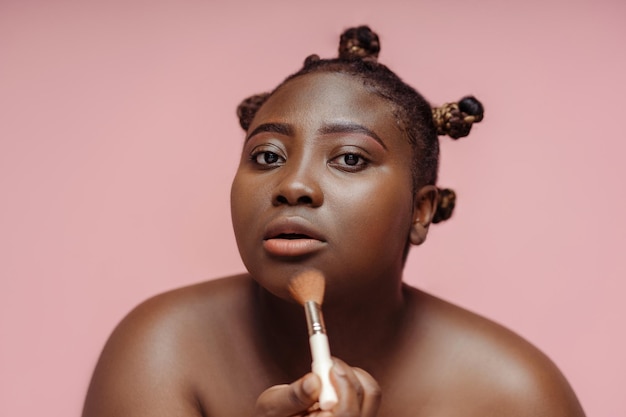 Geconcentreerde Afrikaanse vrouw die make-up maakt met behulp van een make-up kwast die in de spiegel kijkt geïsoleerd op roze achtergrond Beauty concept make-up cosmetica