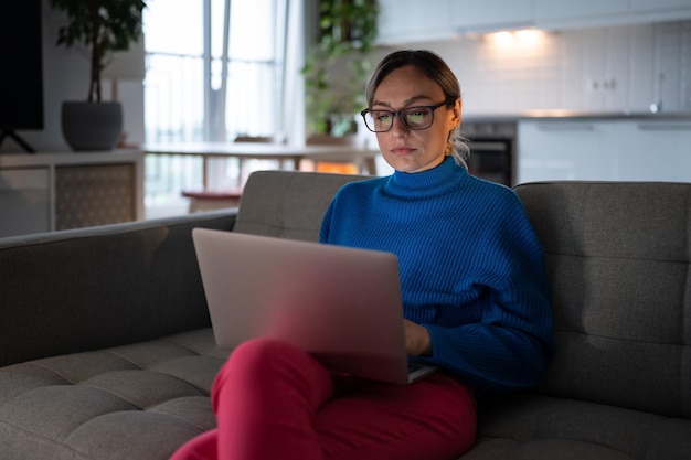 Geconcentreerde Amerikaanse vrouw met een bril die naar het scherm van een laptop kijkt, zit op de bank en werkt thuis online