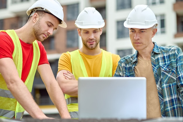 Geconcentreerde ingenieur en twee bouwers in veiligheidshelmen en veiligheidsvesten met behulp van een computer op de bouwplaats