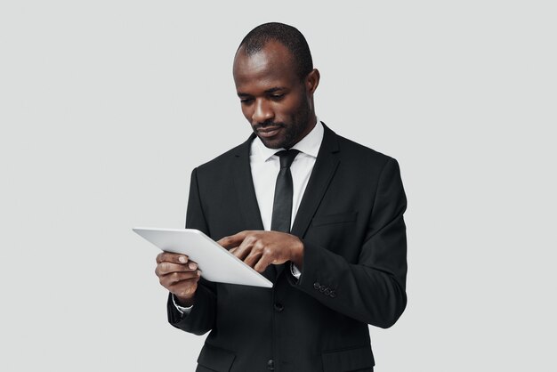 Geconcentreerde jonge Afrikaanse man in formalwear die met digitale tablet werkt terwijl hij tegen een grijze muur staat
