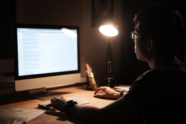 Geconcentreerde jonge aziatische man die een computer gebruikt die thuis in een donkere kamer zit