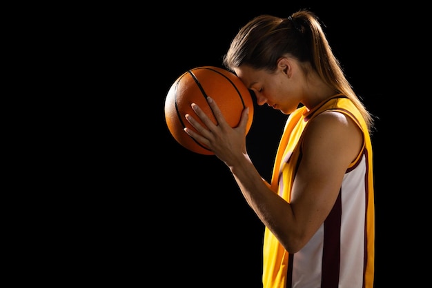 Geconcentreerde jonge blanke vrouwelijke basketbalspeler wiegt een basketbal in een studio op een zwarte