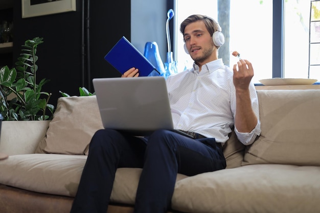 Geconcentreerde jonge freelancer zakenman zittend op de bank met laptop en het onderzoeken van documenten.