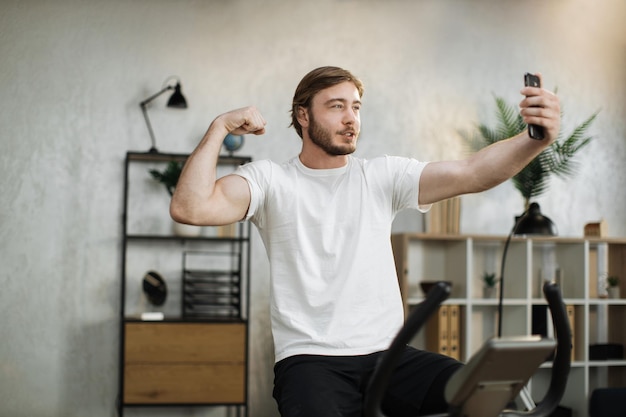 Geconcentreerde jonge man die sportkleding draagt en selfie doet met biceps terwijl hij een hometrainer gebruikt