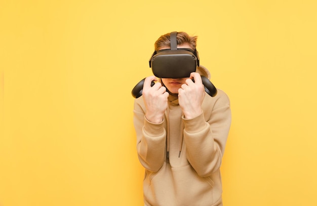 Geconcentreerde jonge man in een VR-helm en controllers in zijn handen staat in een doos op een gele achtergrond en kijkt in de camera Guy vecht in een VR-helm Geïsoleerde VR-gaming