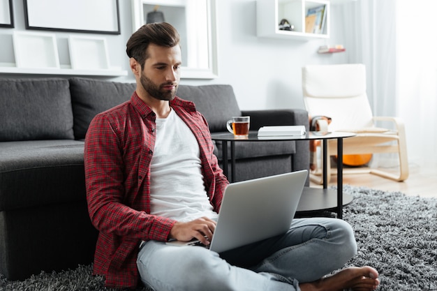 Geconcentreerde jonge man zit binnenshuis met behulp van laptopcomputer.