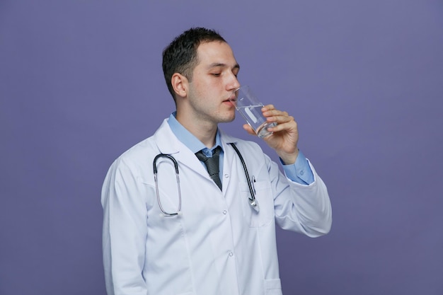 Geconcentreerde jonge mannelijke arts met een medisch gewaad en een stethoscoop om de nek met glas water in de buurt van de mond op zoek naar glas en klaar om het te drinken geïsoleerd op paarse achtergrond