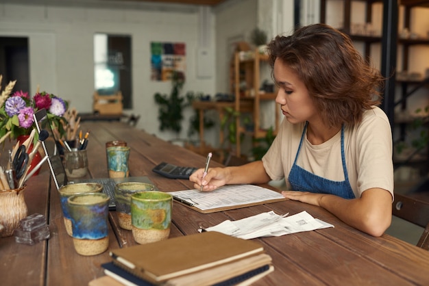 Geconcentreerde jonge vrouw zit aan houten tafel met keramische mokken en analyseert bonnetjes terwijl ze aan het werk is.