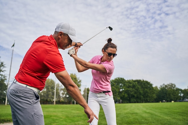 Geconcentreerde jonge vrouwelijke golfspeler die een strijkijzer boven haar hoofd houdt, geholpen door haar persoonlijke trainer