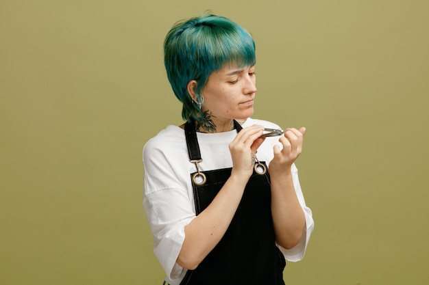 Geconcentreerde jonge vrouwelijke kapper in uniform die haar nagels knipt met nagelknipper geïsoleerd op olijfgroene achtergrond