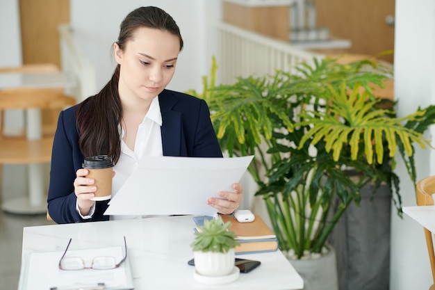 Geconcentreerde jonge zakenvrouw die afhaalkoffie drinkt bij het analyseren van cijfers in het rapport