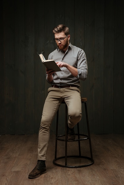 Geconcentreerde knappe jonge, bebaarde man in een bril die zit en leest een boek geïsoleerd op een zwart houten oppervlak