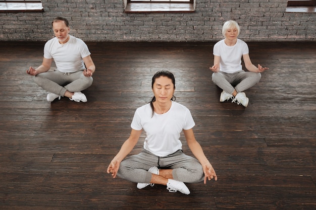 Geconcentreerde langharige trainer die een wit t-shirt en een grijze broek draagt die zijn benen kruist en de armen op de knieën zet terwijl hij in yogapositie zit