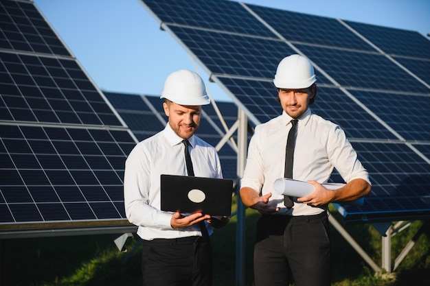 Geconcentreerde mannelijke ingenieurs in hardhats die een laptop gebruiken en een project bespreken terwijl ze naast fotovoltaïsche batterijen op het platteland staan