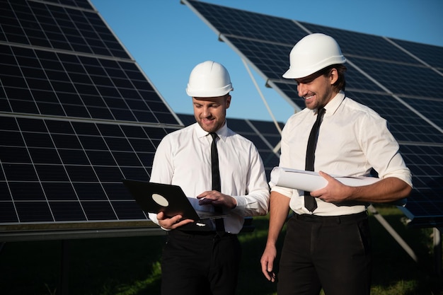 Geconcentreerde mannelijke ingenieurs in hardhats die een laptop gebruiken en een project bespreken terwijl ze naast fotovoltaïsche batterijen op het platteland staan