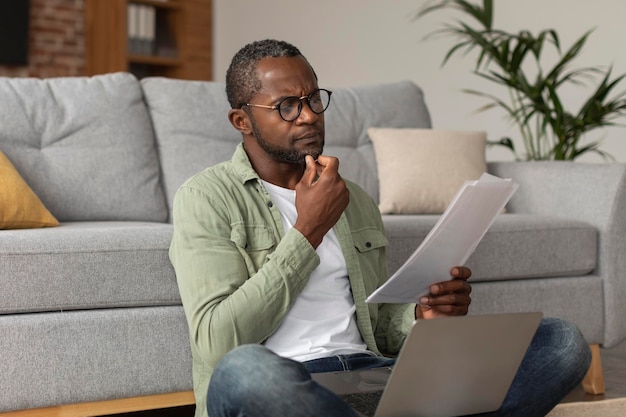 Geconcentreerde peinzende Afro-Amerikaanse man van middelbare leeftijd in glazen met laptop leest document