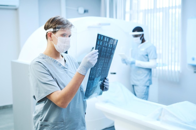 Geconcentreerde radioloog in veiligheidsschild die met hersenröntgenbeeld werkt tegen CT-scanner in het ziekenhuis