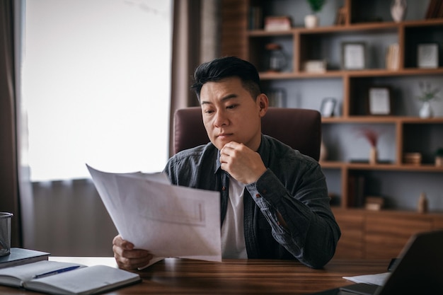 Geconcentreerde rustige chinese man van middelbare leeftijd aan tafel werkt met documenten in het interieur van een thuiskantoor