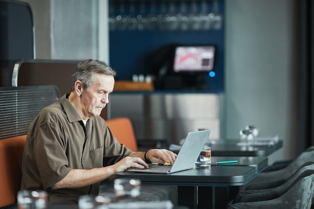 Geconcentreerde senior blanke man met snor aan tafel zitten en werken met laptop in café