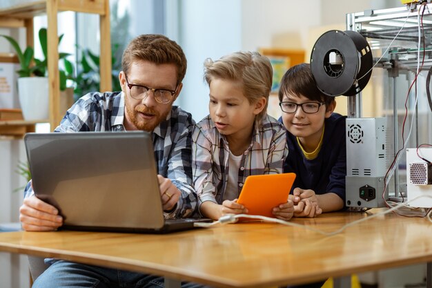 Geconcentreerde tutor die bij zijn studenten zit en laptop aansluit op 3D-printer om te beginnen