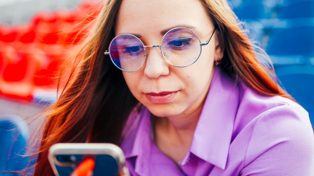 Geconcentreerde volwassen vrouw met lang bruin haar in blouse en bril zittend op de stoel van blauwe tribune en berichten op smartphone