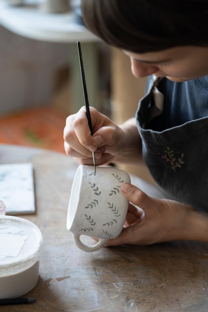 Foto geconcentreerde vrouw pottenbakker zit aan tafel met borstel en minimalistisch patroon schilderen op klei cup