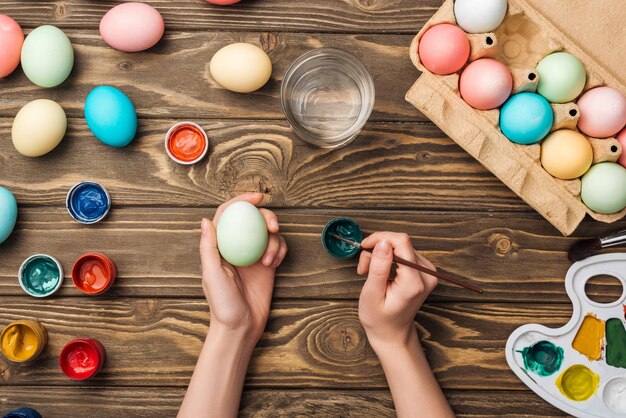 Gedeeltelijke weergave van meisje paaseieren versieren met penseel aan houten tafel met verf en kleur