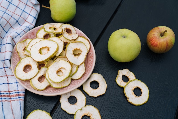 Gedroogde appelschijfjes in open glazen pot. Zelfgemaakte biologische gedroogde appelspaanders met verse appel op zwarte lijstachtergrond. Zoete veganistische snack. Gezond en voedingsconcept. Ondiepe scherptediepte.