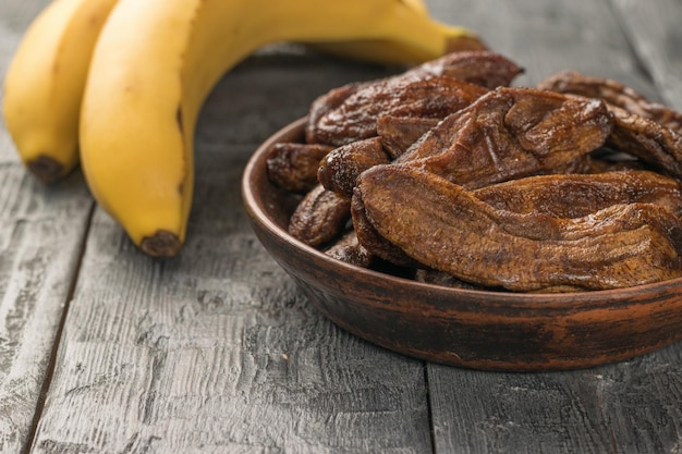 Gedroogde bananen in een kleikom en twee rijpe bananen op een houten tafel Heerlijke natuurlijke fruitsnack