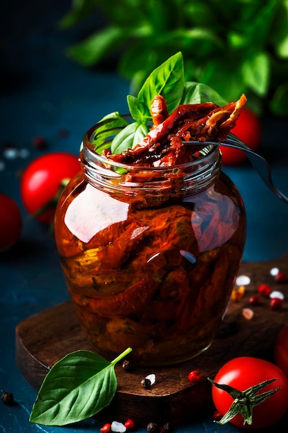 Gedroogde tomaten in olijfolie met groene basilicum en kruiden in glazen pot op blauwe keukentafel voor tekst