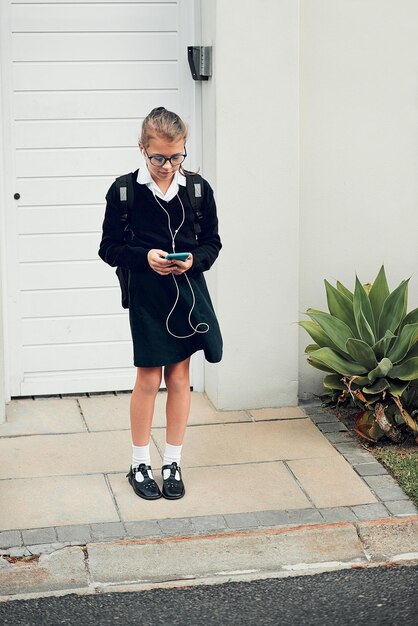 Geduldig wachten op mijn rit naar school om te arriveren Volledige opname van een jong schoolmeisje dat naar muziek luistert en haar mobiele telefoon gebruikt terwijl ze buiten haar huis staat