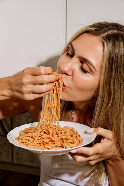 Gedurfde sexy langbenige schoonheid in een witte bodysuit eet spaghetti met tomatenpuree met haar handen Moderne huisvrouw eet pasta in een moderne scandinavische keuken zit op de keukentafel