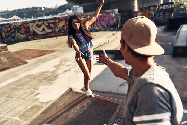 Geef me een hand! Mooie jonge vrouw die naar haar vriend probeert te klimmen terwijl ze buiten tijd doorbrengt in het skatepark