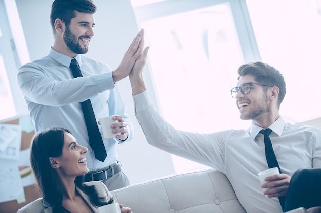 Geef me een high-five! Twee knappe man die high-five geeft en koffiekopjes vasthoudt met een glimlach terwijl hij op de bank op kantoor zit met hun mooie collega