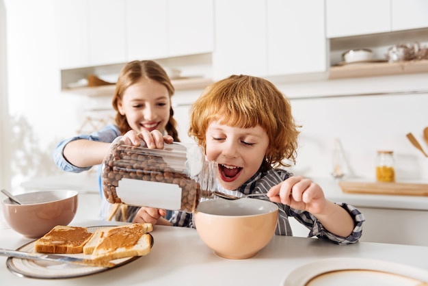 Geef mij meer. Levendige, gepassioneerde vrolijke kinderen die er opgewonden uitzien voordat ze wat ontbijtgranen met chocoladesmaak eten terwijl zijn zus het in zijn kom giet