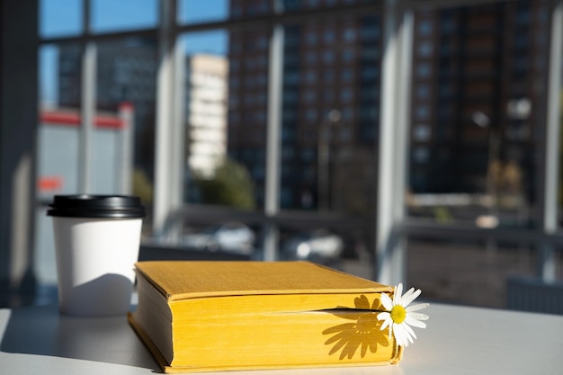 geel boek met kamillebloem en aromatische koffie in een papieren wegwerpbeker ligt op tafel in een café