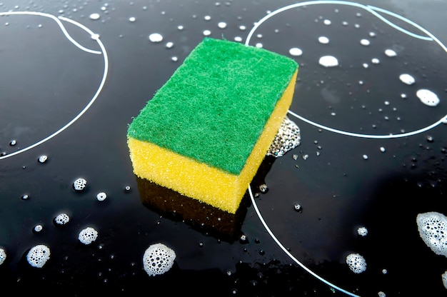 Geelgroene spons op het zwarte oppervlak van de kookplaat en afwasmiddel schuim