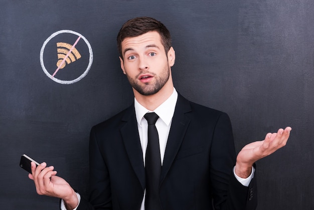 Geen connectie. Gefrustreerde jonge man die telefoon vasthoudt en verbaasd kijkt vanwege een slechte verbinding terwijl hij tegen het delen van symboolkrijttekening op het bord staat