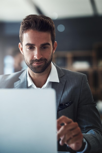 Geen enkele werktaak is te zwaar voor hem. Shot van een knappe jonge zakenman die een laptop gebruikt aan zijn bureau in een modern kantoor