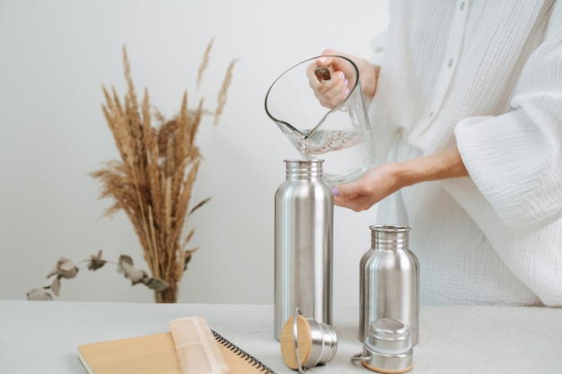 Geen hoofdbeeld van een vrouw die water in een metalen thermoskan uit een kan giet. Midden op een tafel.