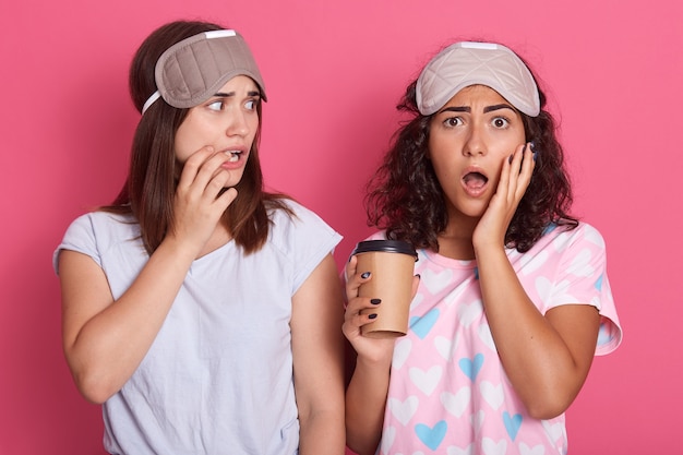 Geërgerd meisje met papieren kopje koffie voor haar wang met palm en poseren in de buurt van haar vriend met wijd geopende mond