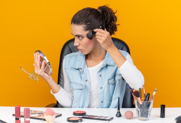 Geërgerd mooie blanke vrouw zittend aan tafel met make-up tools houden spiegel en blozen toe te passen met make-up borstel kijken naar kant geïsoleerd op oranje muur met kopie ruimte