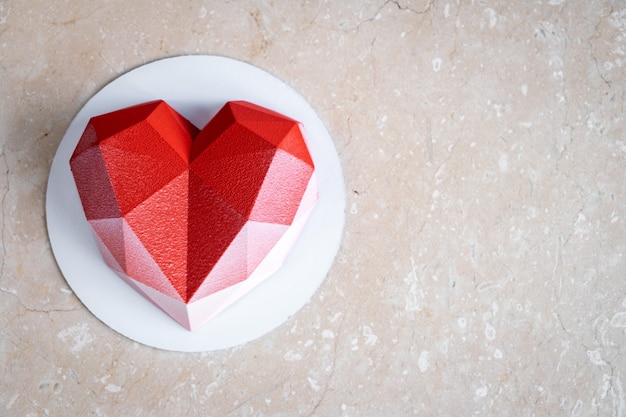 Foto gefacetteerde rood hart mousse cake met velours coating op een zachtroze marmeren tafel.