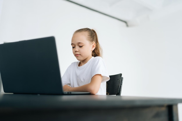 Gefocust blond meisje dat aan een zwarte tafel zit met een laptopcomputer en op zoek is naar online leren op pc elearning film kijken