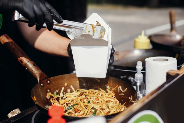 Gefrituurde chinese japanse noedels met groenten en garnalen in afhaaldoos voedselbezorging chef-kok stopt noedels in kartonnen doos om mee te nemen vanuit open keuken aziatisch straatvoedselfestival