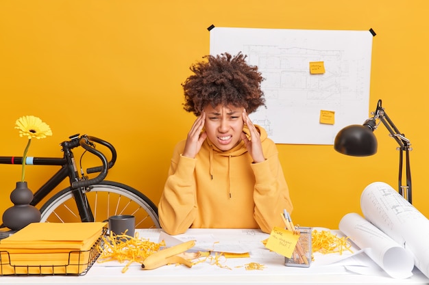 Foto gefrustreerde uitgeputte afro-amerikaanse vrouw raakt tempels aan, lijdt aan ernstige hoofdpijn, moe van het werken met papieren, bereidt architecturaal project voor heeft moeilijke taak