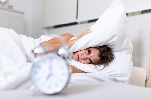 Gefrustreerde vrouw die haar oren bedekt met haar kussen van de wekker in de ochtend. Portret van een geïrriteerde jonge vrouw die oren bedekt met een kussen vanwege de wekker die 's ochtends afgaat, terwijl ze in bed ligt