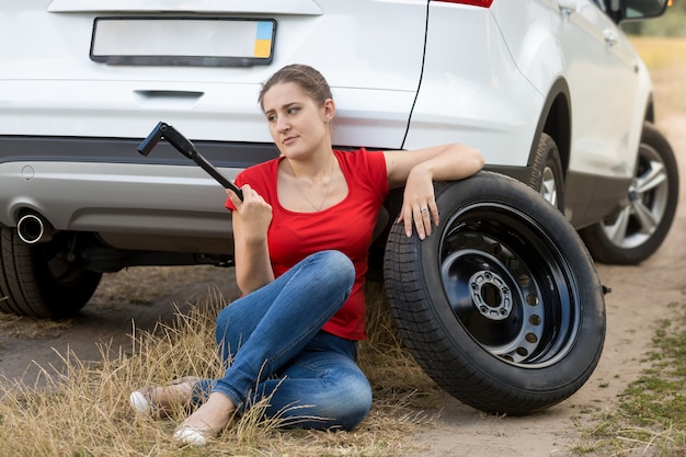 Gefrustreerde vrouw die naast een kapotte auto zit en probeert een lekke band te verwisselen
