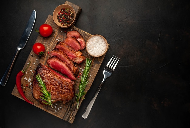 Gegrilde biefstuk, kruiden en specerijen op een snijplank op een stenen achtergrond, bovenaanzicht