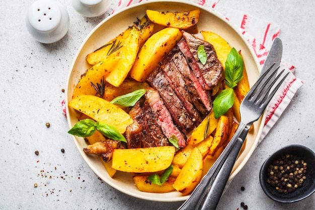 Gegrilde biefstuk met aardappelen en basilicum in een witte plaat, bovenaanzicht.